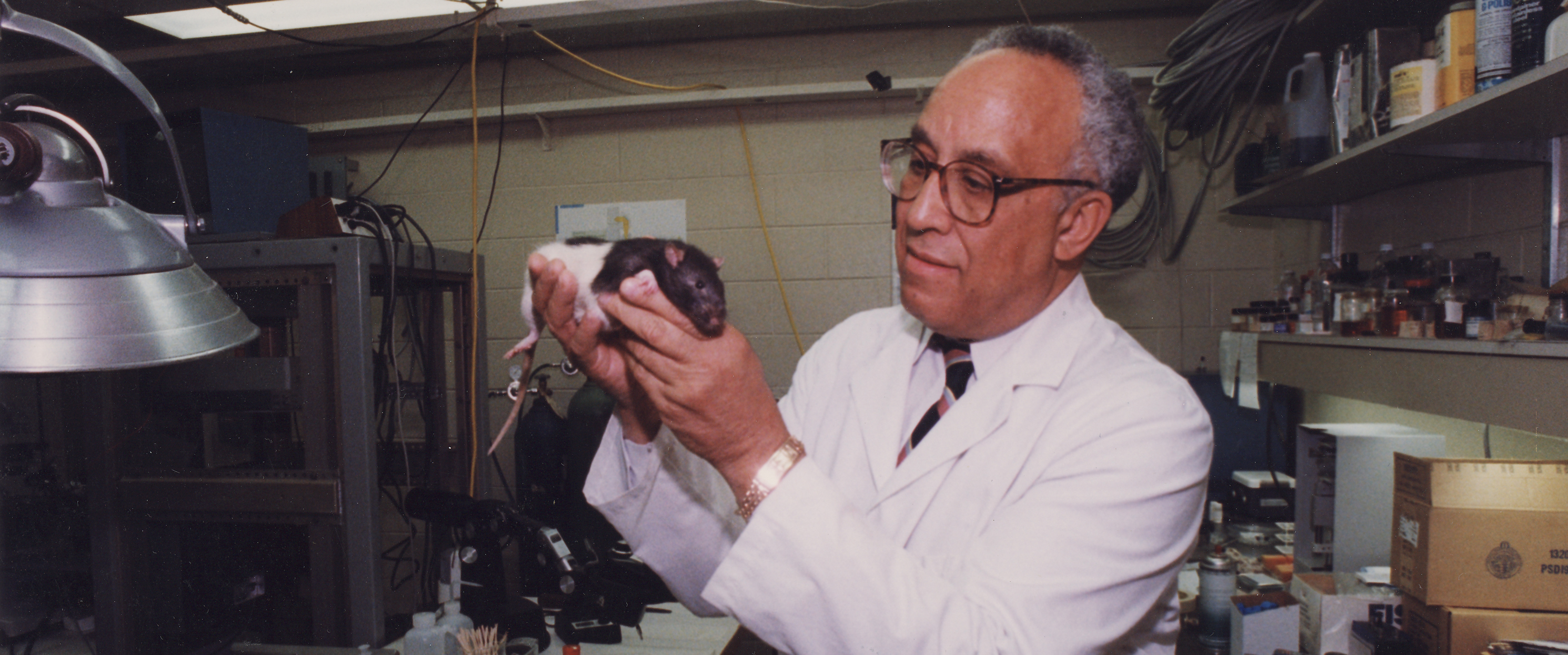 An African American man wearing a lab coat and holding a small white and black rat. He is surrounded by laboratory equipment.